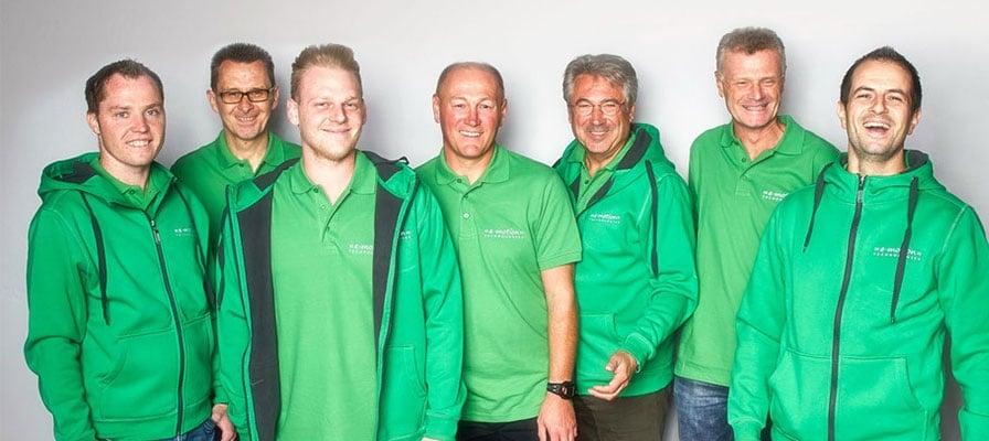 Gruppe von sieben Männern in grünen Oberteilen vor neutralem Hintergrund