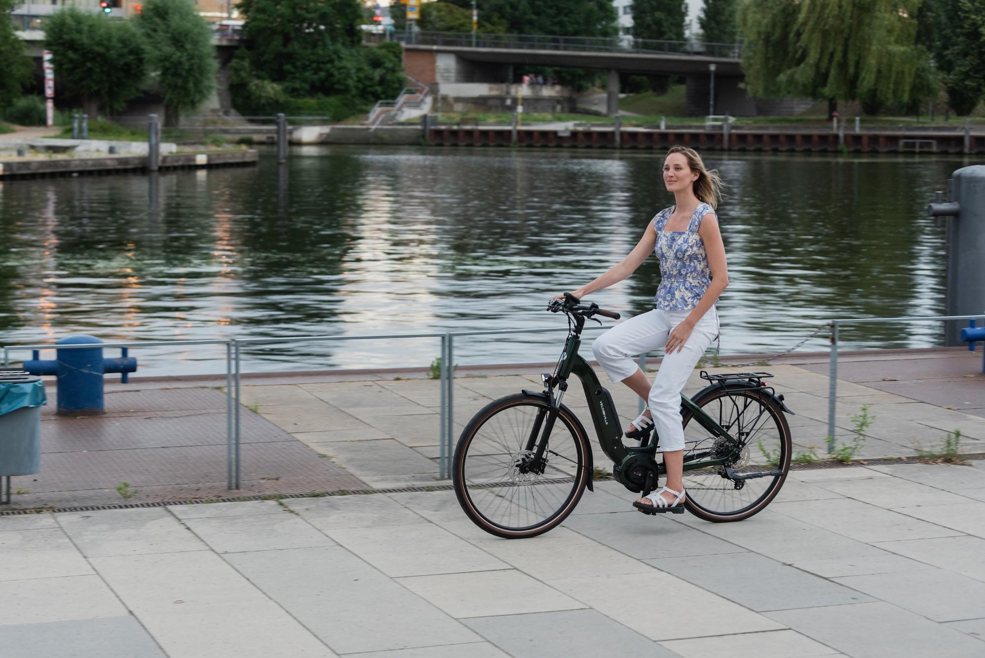Das Bild zeigt eine Frau, die auf einem E-Bike der Marke Velo de Ville fährt. Das Fahrrad ist mit einem modernen Ra