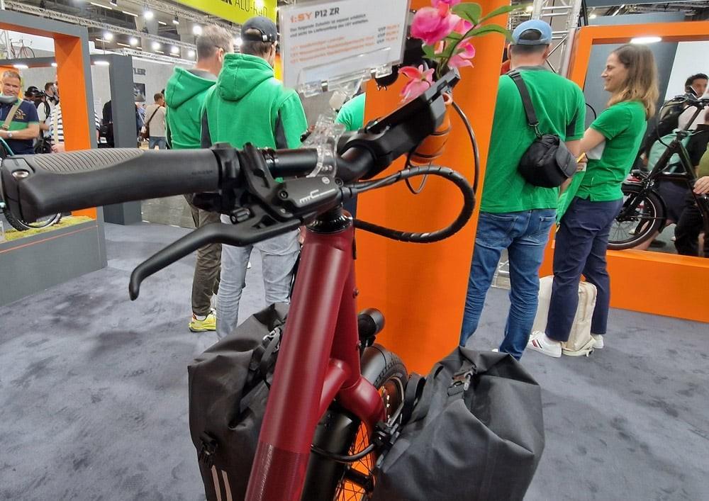 Detail eines e-Bikes mit Lenker, ergonomischen Griffen und farbigen Blüten, im Hintergrund interessierte Besucher.
