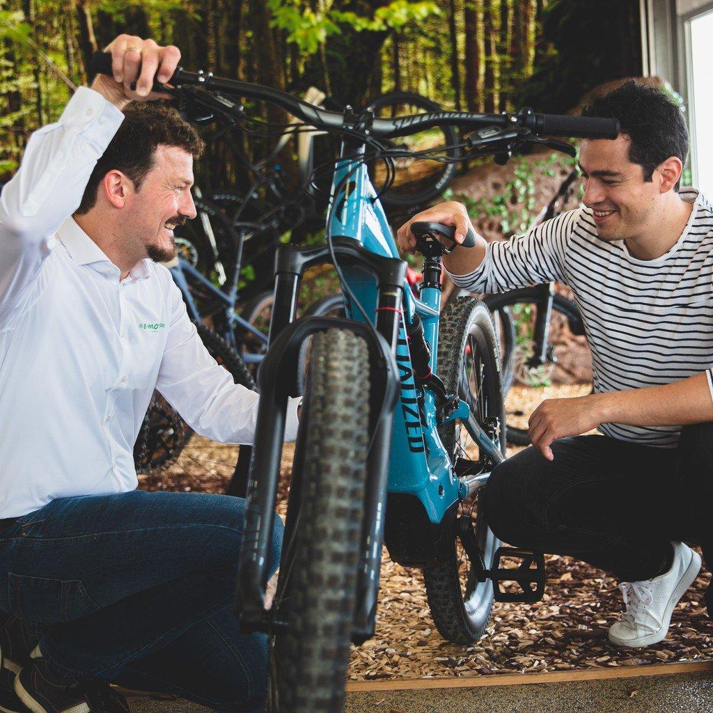 Zwei Männer sind in einem Fahrradgeschäft. Der Mann links trägt ein weißes Hemd und zeigt einem Mann in einem gestr