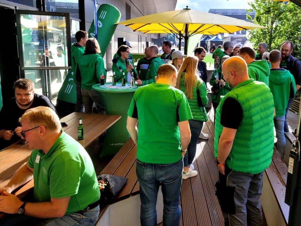 e-motion Mitarbeiter in grüner Kleidung an Stehtisch mit Getränken und sonniger Szene auf der Eurobike 2025.