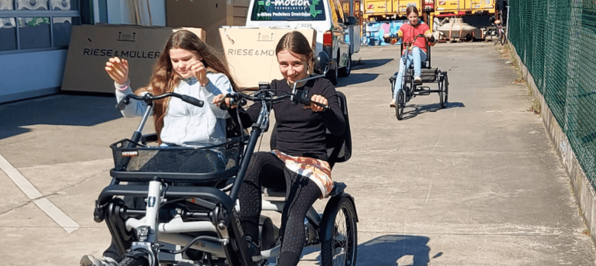 Zwei Mädchen fahren lachend ein Dreirad mit Frontsitz, ein weiteres Kind folgt auf einem eigenen Dreirad.