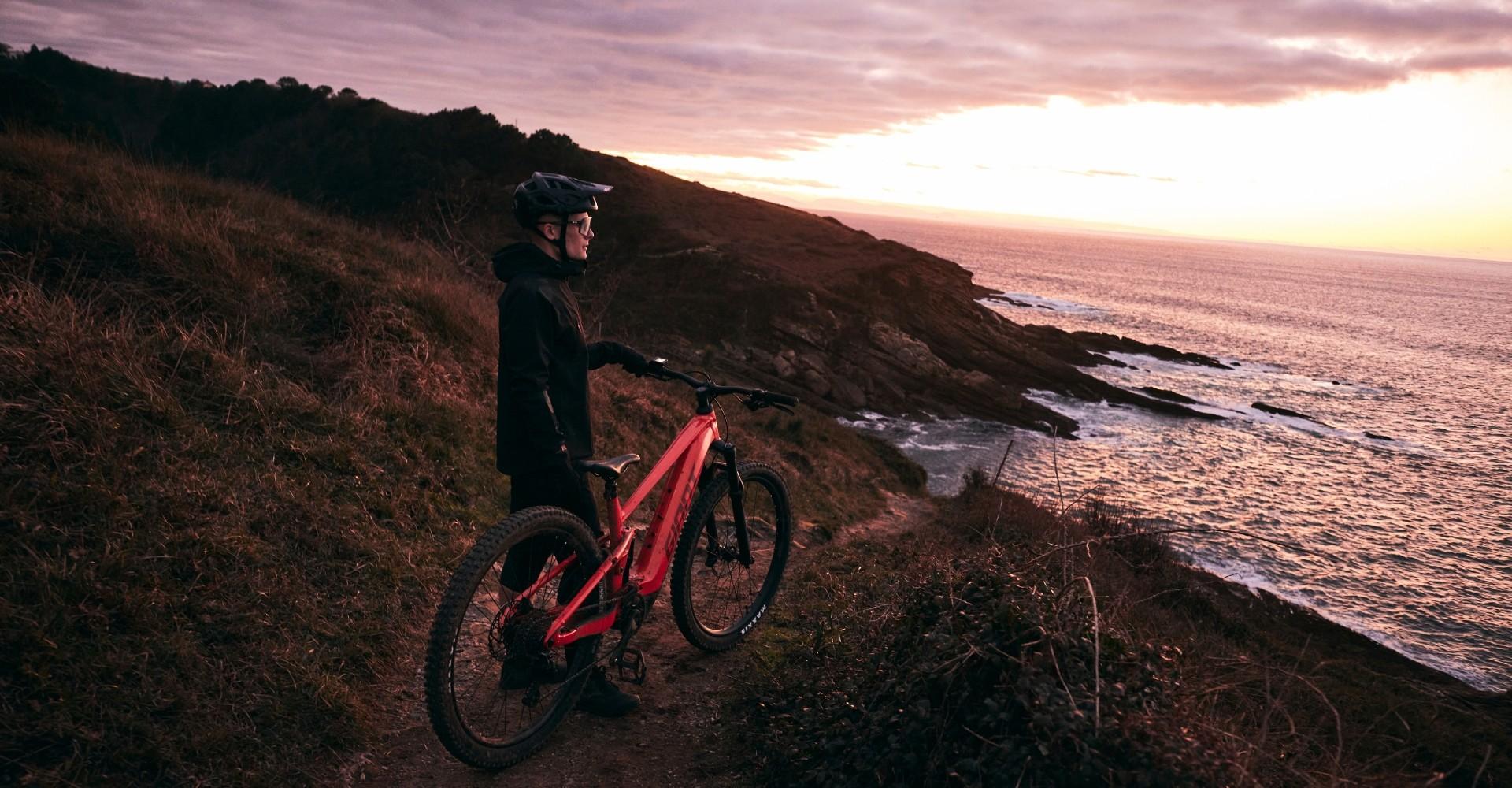 Ein Radfahrer mit rotem Giant Stance E+ steht auf einem Küstenpfad und blickt beim Sonnenuntergang aufs Meer.