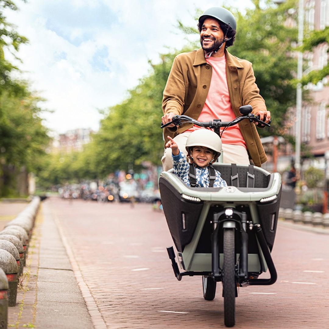 Ein Mann fährt mit einem Lastenrad durch die Stadt, vorne in der Transportbox sitzt ein Junge