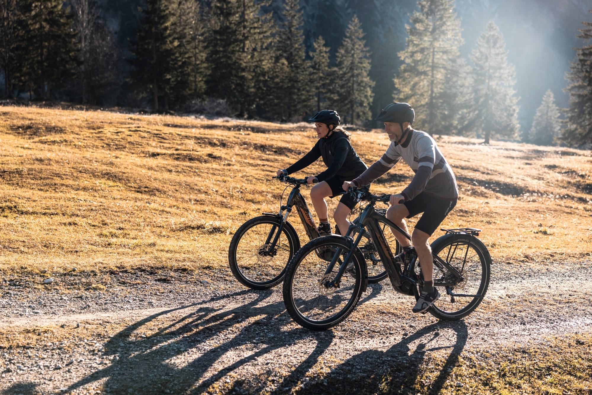 Das Bild zeigt zwei Männer in sportlichen Outfits, die mit einem e-MTB einen Berg hochfahren.