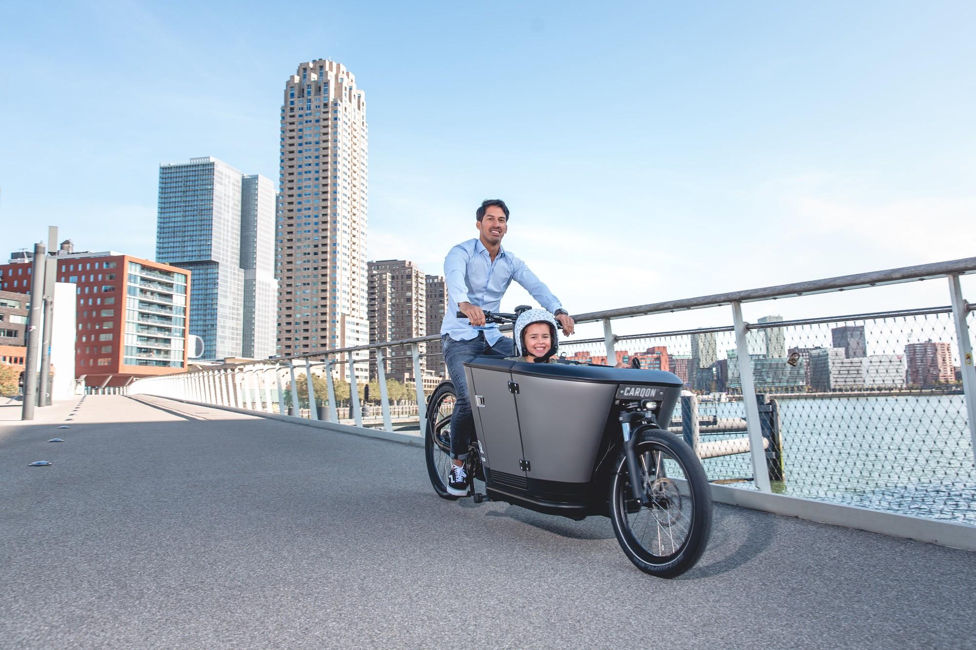 Das Bild zeigt ein Carqon E-Lastenrad, das von einem Mann in einer städtischen Umgebung gefahren wird.