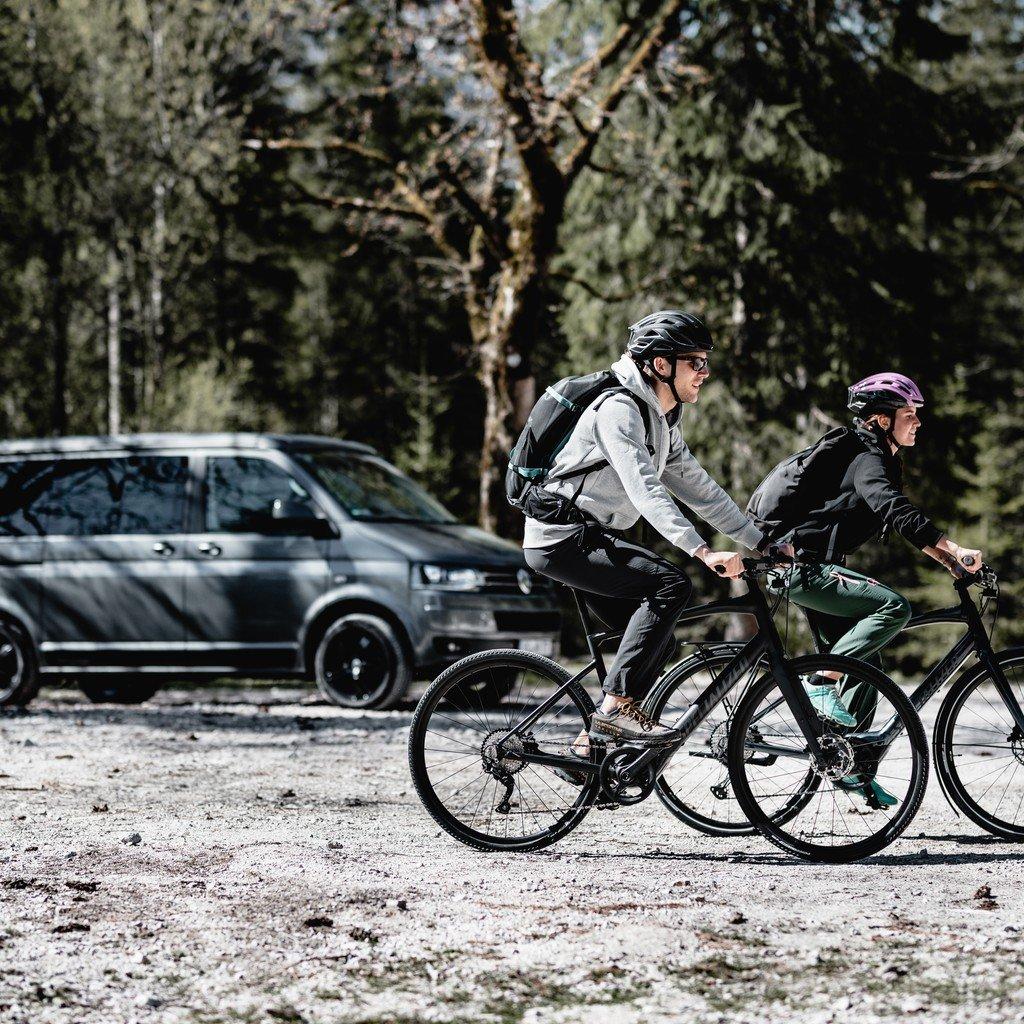 Zwei Radfahrer tragen Helme und fahren nebeneinander auf einem Fahrradweg durch einen Wald. Im Hintergrund steht ei