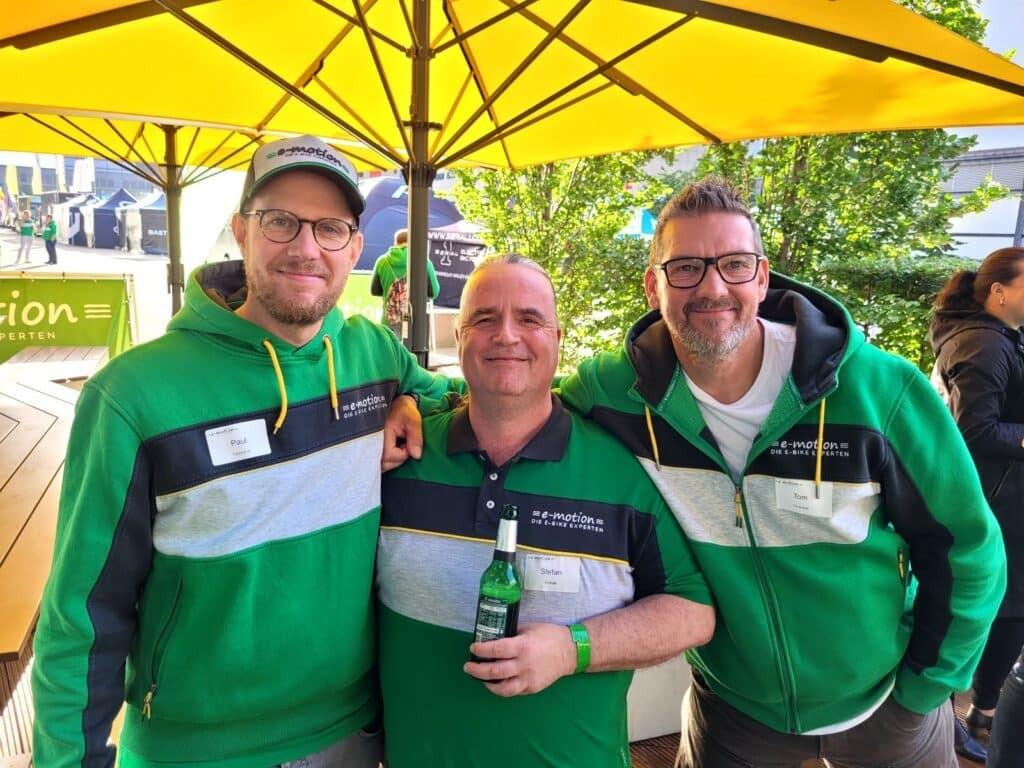 Drei Männer in grünen e-motion Sweatshirts unter gelbem Sonnenschirm, entspannte Messeatmosphäre.