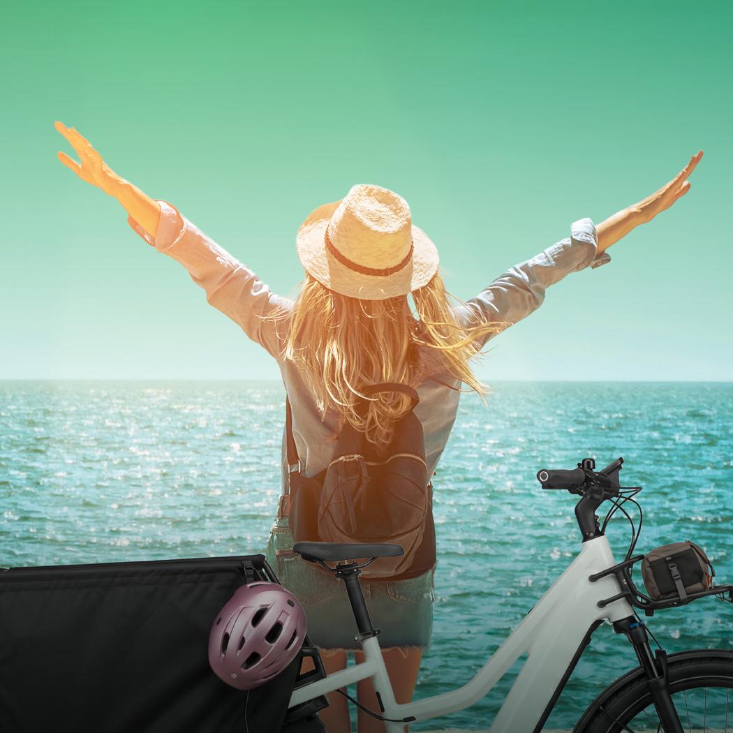 Eine Frau steht vor dem Wasser und breitet die Arme aus, hinter ihr ein e-Bike