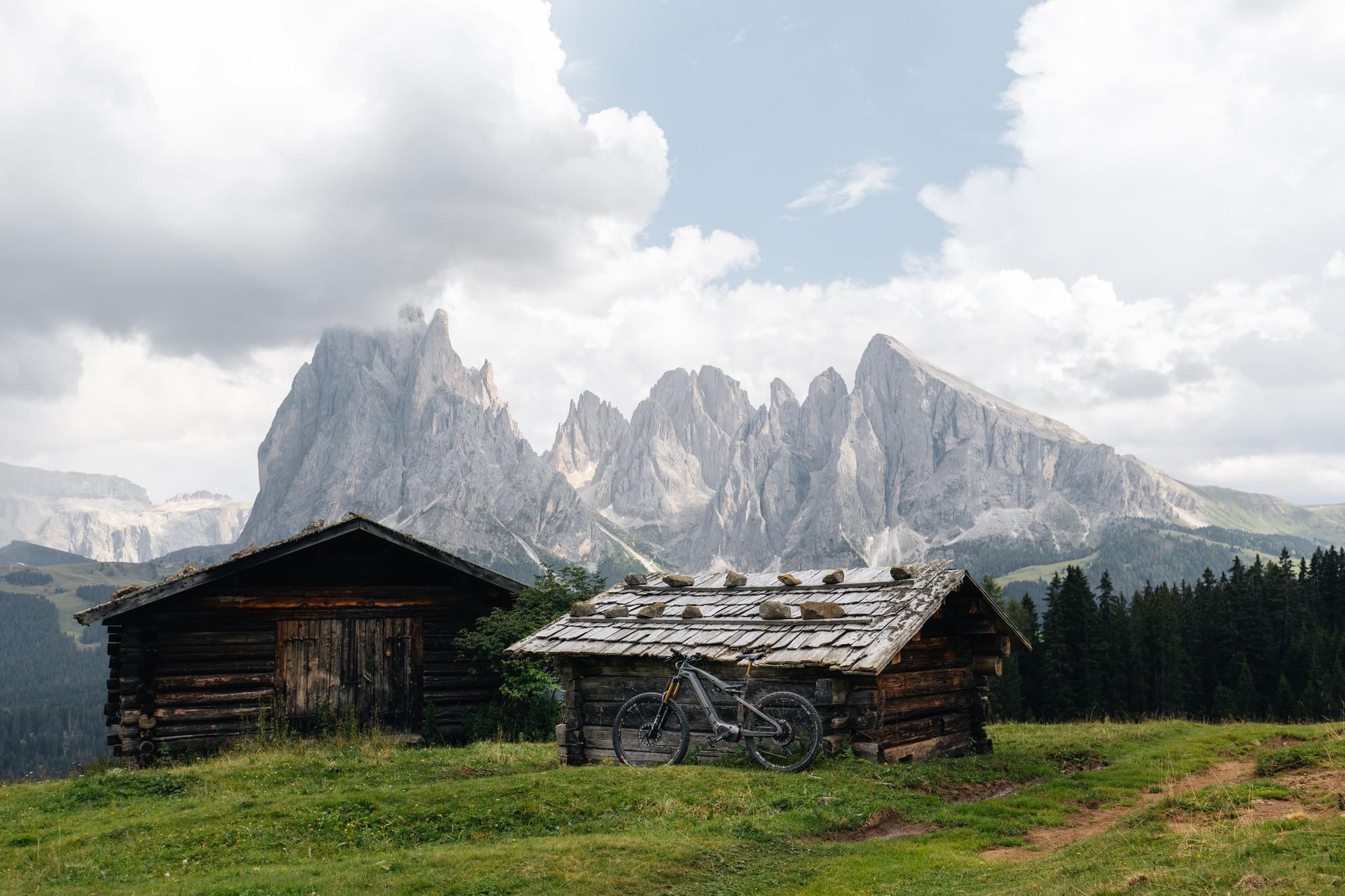 Das Bild zeigt ein E-Bike, das vor zwei Holzhütten in einer beeindruckenden Berglandschaft positioniert ist.