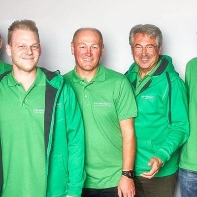 Gruppe von sieben Männern in grünen Oberteilen vor neutralem Hintergrund