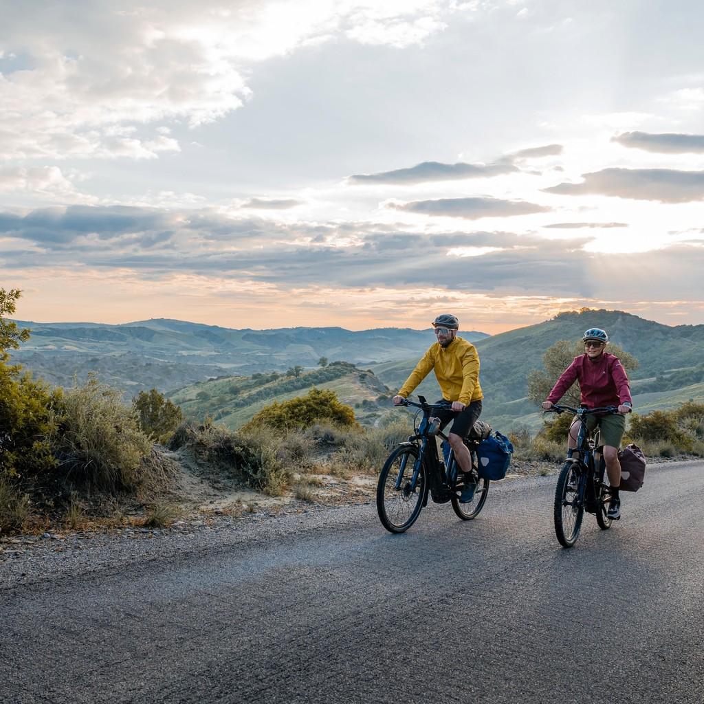 Zwei Personen auf Bosch Performance Line E-Bikes fahren bei Sonnenuntergang durch eine malerische Landschaft.