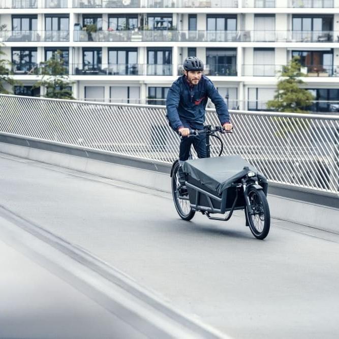 Mann fährt Riese & Müller Lastenrad auf Brücke vor modernen Gebäuden