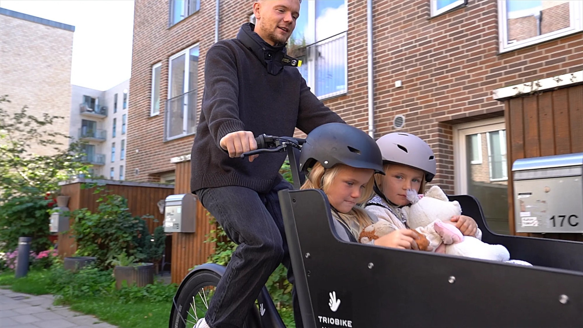 Ein Mann fährt mit dem Triobike Hafnia Lastenrad und zwei Kindern in der Transportbox durch die Stadt