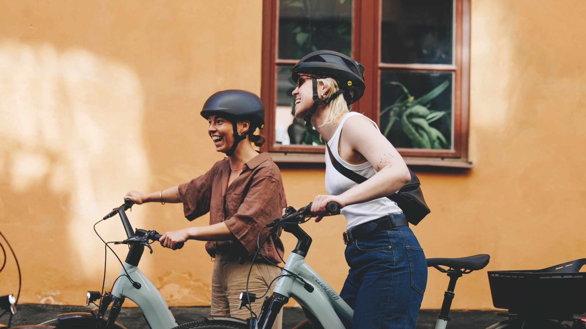 Zwei Frauen fahren freudig Specialized Como E-Bikes vor einem Gebäude. Die stilvollen E-Bikes bieten Komfort und El