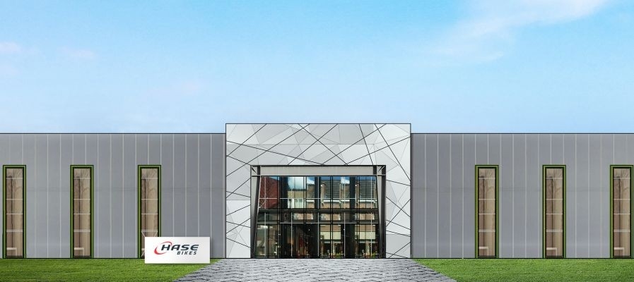 Moderne Halle mit grauer Fassade und großen Fenstern, Logo von Hase Bikes im Vordergrund, Grünfläche davor.