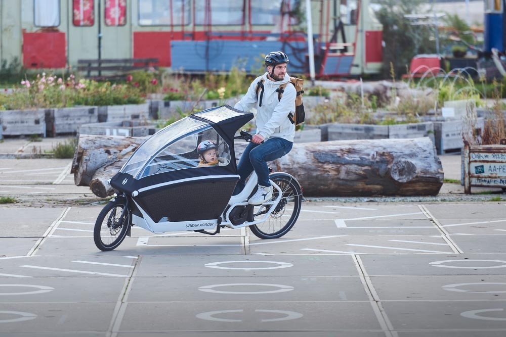 Ein Mann fährt mit Kind in der Transportbox im Urban Arrow Lastenrad mit Regenschutz